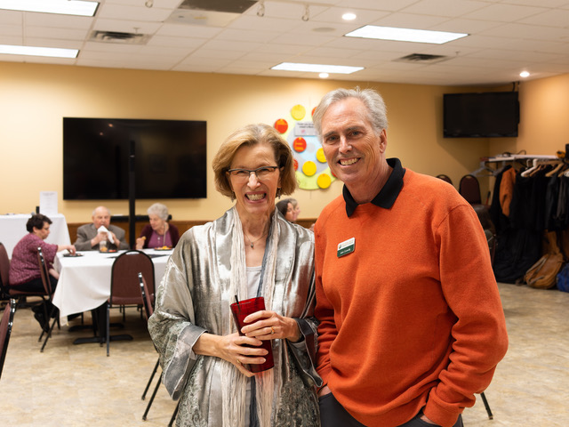 Shirley Barnes is happy to greet John Connolly, Twin Cities North Chamber of Commerce. Description: Women and man standing next to each other. The man is wearing an orange sweater and the woman is wearing a gray silver cardigan.