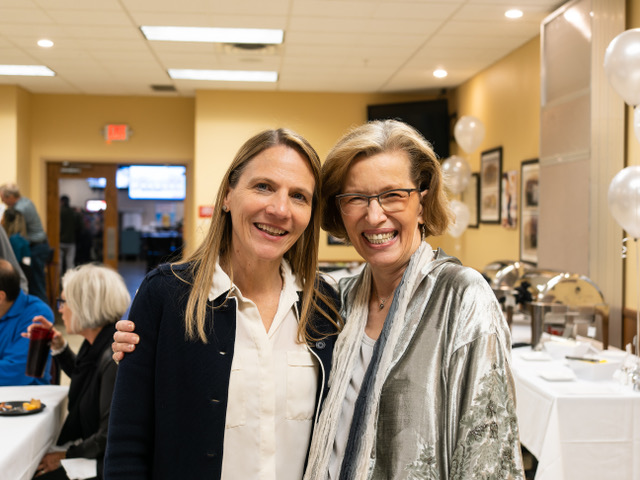 Shirley Barnes is happy to celebrate with local businesses.  Description: Two women face the camera smiling. The Woman on the left is wearing a black dress jacket, while the woman on the right is wearing a gray cardigan.